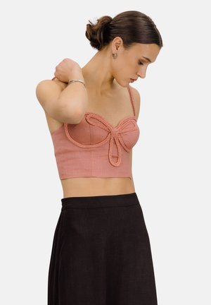 Ung kvinne med mørkt hår i en knute, iført en korallfarget crop-top med flettet detaljering og et høytlivsskjørt i svart, mot en ensfarget bakgrunn.