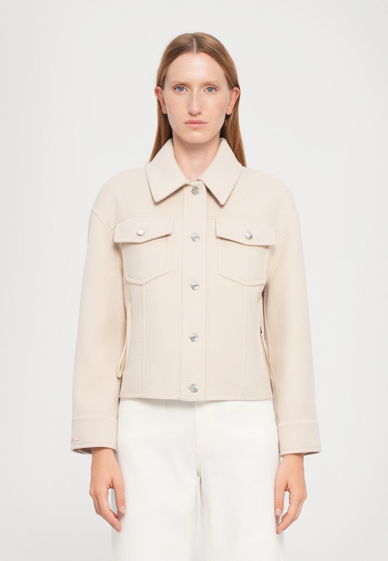 Giacca cropped beige in tessuto morbido, dotata di colletto, bottoni argentati, due tasche sul petto e leggere fessure laterali nei polsini.