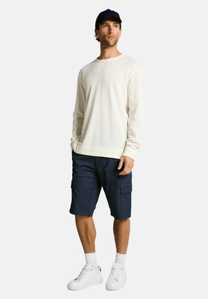 Mann mit schwarzer Kappe, weißem langärmligem Hemd, marineblauen Cargoshorts, weißen Socken und weißen Sneakers steht mit den Händen in den Taschen.