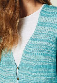 Une femme portant un t-shirt blanc à col rond sous un cardigan en tricot à carreaux bleu et blanc avec des boutons, avec de longs cheveux châtains clairs reposant sur son épaule.
