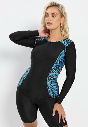 Mujer con camiseta de baño de manga larga negra con paneles de estampado de leopardo azul y shorts de baño negros a juego, posando con una mano en el cuello.