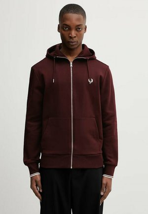 Man draagt een bordeauxrode hoodie met ritssluiting, voorzakken en een klein wit logo, tegen een effen lichte achtergrond.
