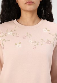 Lys rosa hettegenser med rund hals, prydet med delikat blomsterbroderi i hvit og rosa, med grønne blader som skaper et teksturert design.