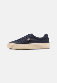 Navy canvas sneaker with waxed laces, featuring a woven jute platform sole and gold logo accent on the side. Smooth texture, round toe design.