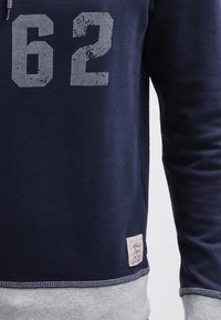 Felpa blu navy con una stampa grigia sbiadita "62", con orlo inferiore grigio e etichetta laterale. Tessuto morbido.