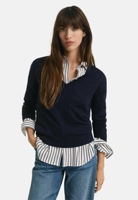 Maritieme gebreide trui met een V-hals over een wit shirt met verticale zwarte strepen. Lijkt gelaagd te zijn met denim jeans.