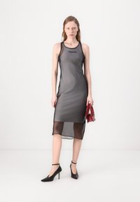 Graues halbtransparentes Kleid mit geradem Schnitt und schwarzem Saum. Verfügt über ein Logo auf der Brust und wird mit schwarzen high heels und einer roten Tasche kombiniert.