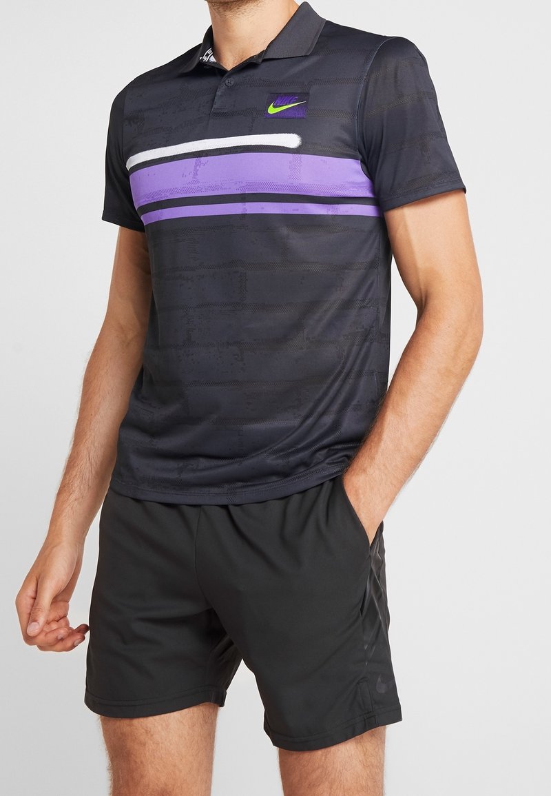 Homme portant un polo noir avec des rayures horizontales violettes et blanches et un short de sport noir, debout avec une main dans la poche.