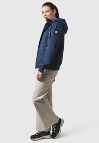 Marinblå jacka med huva, framdragkedja och sidofickor. Kombinerad med ljusbeiga byxor och mörka sneakers. Slät textur, avslappnad design.