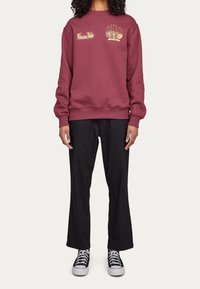 Sudadera color marrón burdeos con diseños gráficos dorados en el pecho, combinada con pantalones holgados negros y zapatillas negras. Tejido texturizado y puños acanalados.