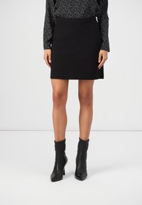 Jupe noire à coupe ajustée, en tissu lisse, avec une ceinture à la taille et deux poches latérales, associée à des bottines noires.