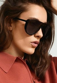 Black oversized sunglasses with a geometric shape, featuring dark lenses and sleek, thin black frames. Textured brown clothing in the background.