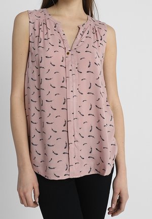 Femme portant un chemisier rose sans manches avec un motif de plumes noires et un seul bouton près de l'encolure, associé à un pantalon noir.