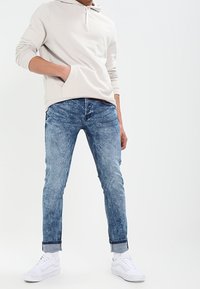 Felpa grigia chiara con tasca frontale, abbinata a jeans blu aderenti e sneakers bianche. I jeans sono leggermente strappati con orli arrotolati.