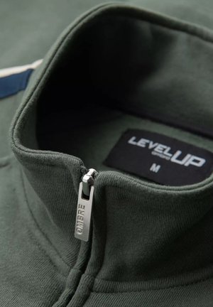 Sudadera verde con cremallera, de alto cuello, que presenta un tirador de metal marcado con "OMBRÉ" y una etiqueta negra que dice "LEVELUP", tamaño M.