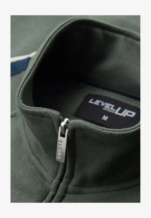 Felpa verde con zip e colletto alto, dotata di un cursore metallico sulla zip contrassegnato con "OMBRÉ" e di un'etichetta nera con la scritta "LEVELUP", taglia M.