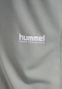 Lys grøn, struktureret stof med et ribbet mønster prydes af en hvid "hummel PERFORMANCE" logo. Formen er flad og i nærbillede.