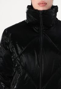 Veste noire matelassée avec un col haut, une fermeture éclair sur le devant et une texture brillante, en relief avec des motifs en forme de diamant.