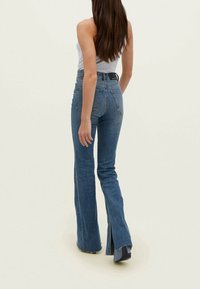 Jeans de mezclilla acampanados con cintura alta, color azul claro y dobladillo sin terminar. Presenta dos bolsillos traseros y una etiqueta de marca sutil en la cinturilla.