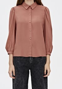 Rose-colored blouse with a classic collar, puffed sleeves, and five button closure. Smooth fabric with minimal sheen. Paired with dark jeans.