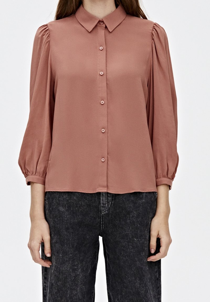 Rose-colored blouse with a classic collar, puffed sleeves, and five button closure. Smooth fabric with minimal sheen. Paired with dark jeans.