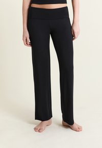 Femme portant un pantalon ample noir et un haut court assorti, debout pieds nus sur un sol clair devant un fond uni.