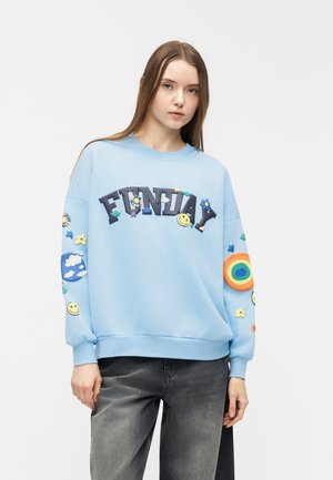 Ung kvinde iført lys blå sweatshirt med teksten "FUNDAY", smileys, skyer, blomster og regnbue-patches, kombineret med falmede sorte jeans.