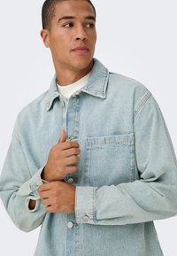 Jasnoniebieska kurtka jeansowa z zapinanym na guziki przodem, spiczastym kołnierzem i kieszenią na piersi. Charakteryzuje się podwiniętymi mankietami z ozdobnymi guzikami oraz luźnym krojem.