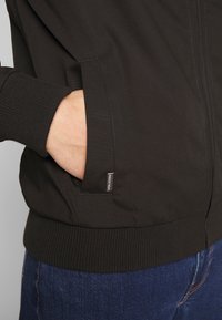 Chaqueta bomber negra con puños y dobladillo acanalados, con bolsillos laterales. El material parece suave, con un brillo sutil y una pequeña etiqueta de marca visible.