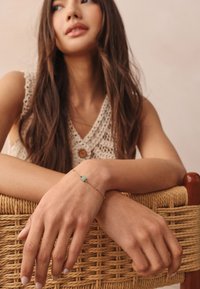 Silver bracelet with a rectangular turquoise stone, featuring a fine chain and small gold accents. Hands resting on a woven basket.