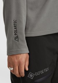 Grijze Polartec fleece mouw met logo, met een gladde textuur. Hand in zwarte Gore-Tex broek, met naaddetails en logo. 