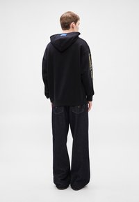 Μαύρο φούτερ με κουκούλα και κορδόνι, με πλευρική σήμανση, συνδυασμένο με φαρδιά σκούρα τζιν παντελόνια με πίσω τσέπες και ανοιχτόχρωμα ράμματα.