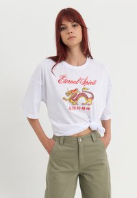 Hvid oversized T-shirt med rund halsudskæring, som prydes af et farverigt dragegrafik og teksten "Eternal Spirit". Knyttet i taljen.