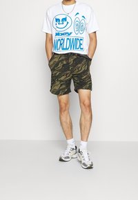 T-shirt blanche avec des motifs graphiques bleus, associée à un short à motifs camouflage en vert et marron. Baskets en caoutchouc avec des accents noirs et jaunes.