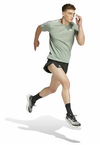 Coureur masculin en maillot vert, short noir et chaussettes noires, en plein élan, courant en avant avec une expression concentrée sur fond blanc.