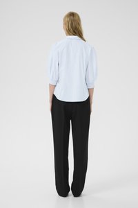 Una persona bionda sta in piedi con le spalle rivolte, indossando una blusa azzurra a maniche a sbuffo con maniche a polsino e pantaloni neri dritti.