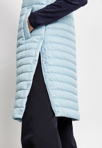 Jasnoniebieska pikowana kamizelka z przeszyciami, bocznymi kieszeniami z rozcięciami, zatrzaskami i gładką teksturą. Nosi się ją na ciemnych spodniach.