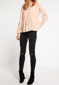 Vaaleanpunainen pleatattu paita, jossa on nappisuljenta ja pitkät hihat, yhdistettynä kuluneisiin mustiin skinny-farkkuihin ja mustiin korkokenkäisiin nilkkureihin.