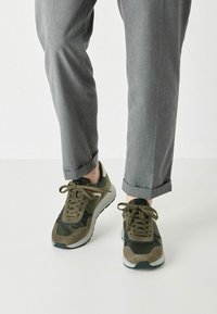 Zapatillas deportivas de color verde oliva con materiales de ante y malla, que presentan una puntera redondeada, diseño de cordones y una suela de goma blanca.
