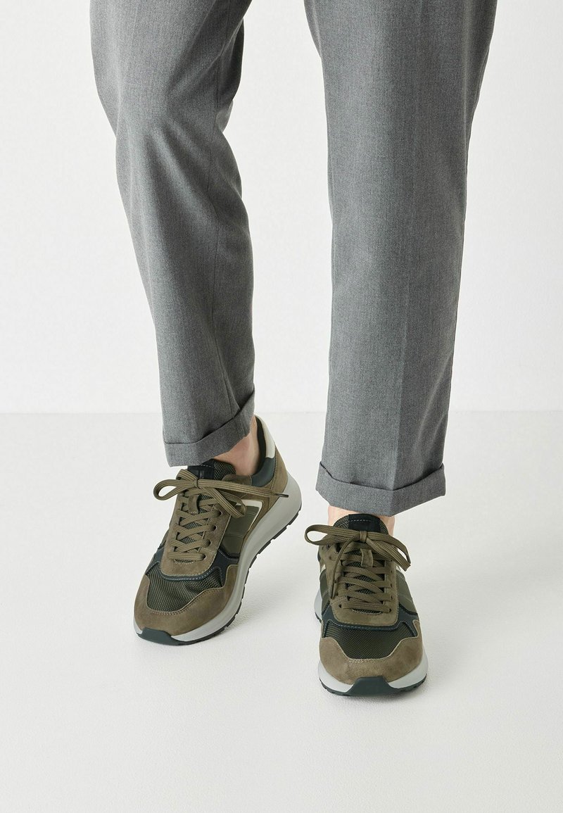 Zapatillas deportivas de color verde oliva con materiales de ante y malla, que presentan una puntera redondeada, diseño de cordones y una suela de goma blanca.