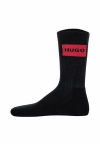 Schwarze Baumwollsocke mit gerippter Struktur, die mit einem auffälligen roten rechteckigen Patch versehen ist, auf dem das Wort "HUGO" in schwarzer Schrift steht.