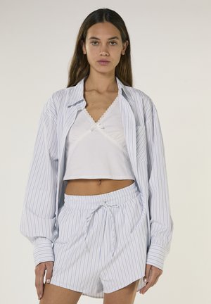Kvinde set forfra iført en hvid cropped camisole, en oversized blåstribet skjorte med knapper og matchende shorts med snor i taljen.