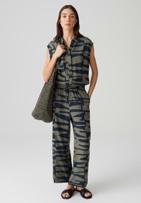 Ärmelloses, knöpfbares Hemd mit grünem und marineblauem abstraktem Muster, kombiniert mit passenden weit geschnittenen Hosen. Das Model hält eine strukturierte graue Tasche und trägt schwarze Sandalen.