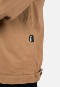 Chaqueta de lona marrón con una textura suave, que presenta una etiqueta lateral, detalles con botones y un corte ligeramente entallado.