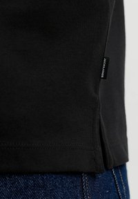 Ourlet de chemise en tissu noir avec fente latérale et petite étiquette noire portant la mention "JACK & JONES" sur un jean en denim bleu.