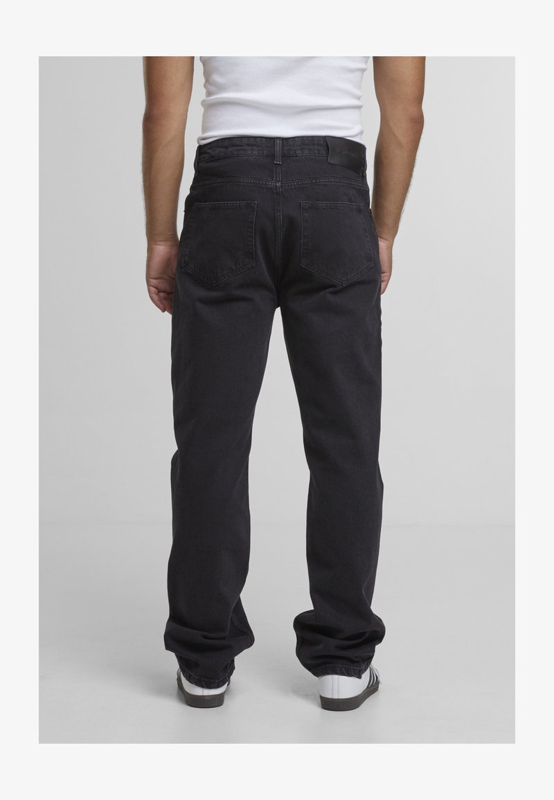 Mann trägt lockere schwarze Jeans und weiße Turnschuhe, steht mit dem Rücken zur Kamera vor einem einfarbigen hellgrauen Hintergrund.