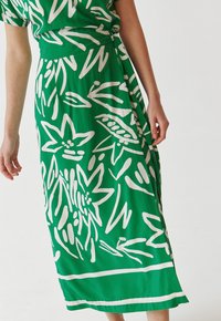 Grüne Skirt mit weißen botanischen Mustern, mit einem fließenden Design und knöchellang. Der Stoff hat eine glatte, leichte Textur.
