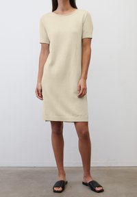 Abito in maglia beige, corto e finto a maniche corte, con scollatura rotonda e orlo a coste. Indossato con sandali neri piatti su uno sfondo neutro.