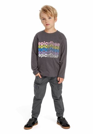 Chemise grise à manches longues avec le texte "vibes épiques" en polices colorées, associée à un pantalon cargo gris avec des poches latérales et des baskets noires.