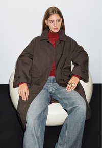 Brauner übergroßer Mantel mit Knopfverschlüssen und seitlichen Taschen, kombiniert mit einem roten Rollkragenpullover und weitgeschnittenen Jeans. Glatte Stofftextur.